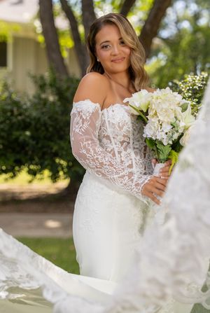Bride in off-shoulder lace wedding dress holding white floral bouquet outdoors near trees.
