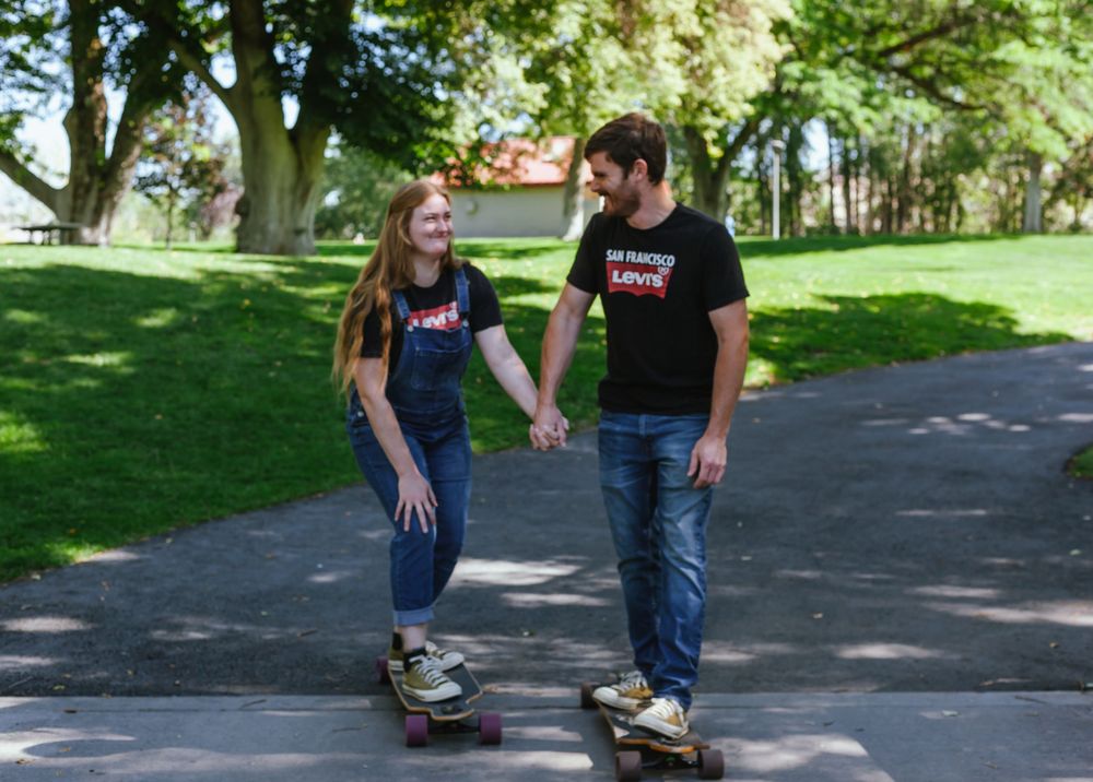 Two people wearing jeans and black t-shirts skateboarding together on a paved path in a sunny park.