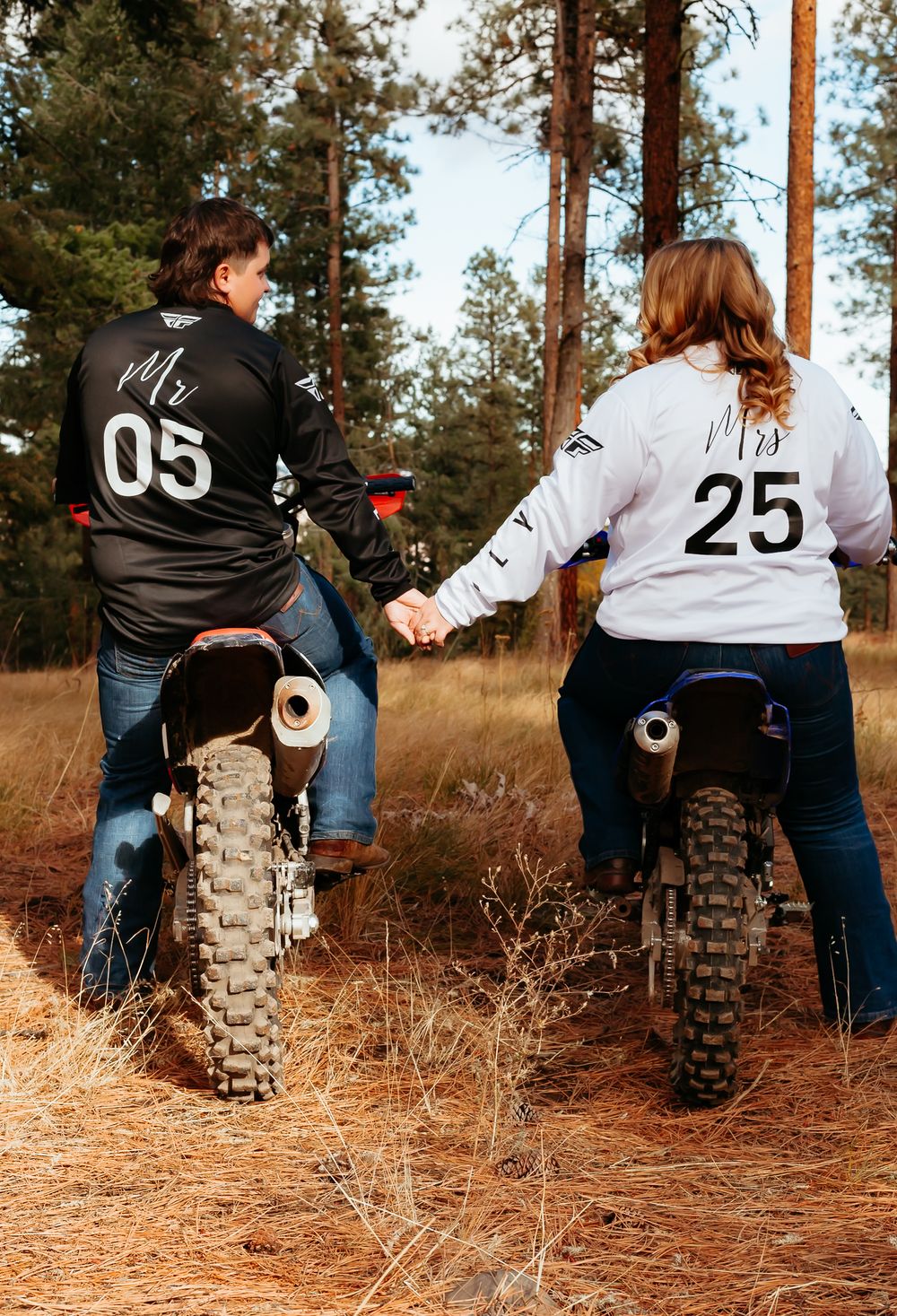 Two people sit on matching dirt bikes wearing numbered jerseys 05 and 25 in a pine forest setting.