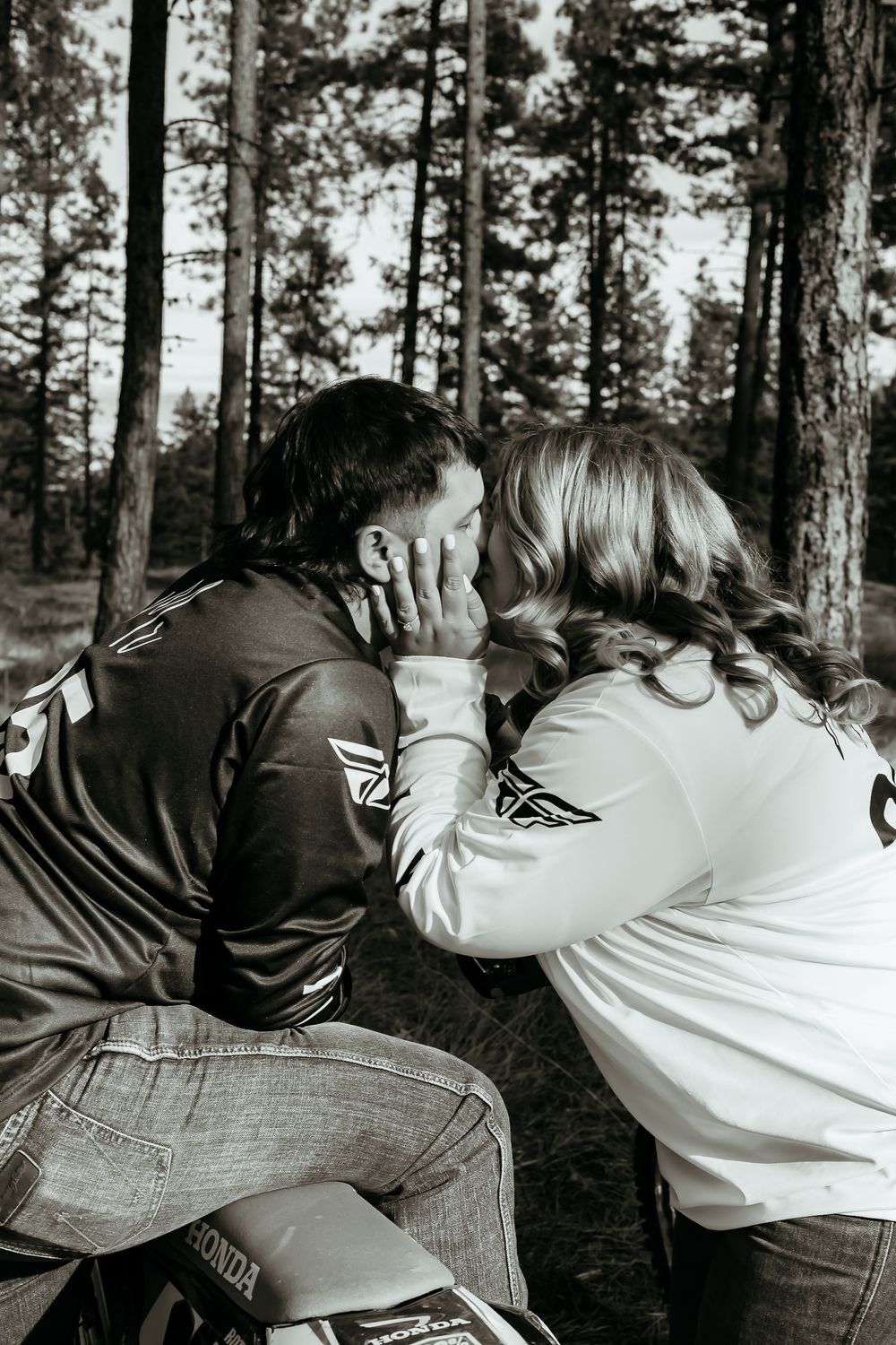 A couple sitting among tall pine trees in the forest while wearing matching varsity jackets on a black and white photo.