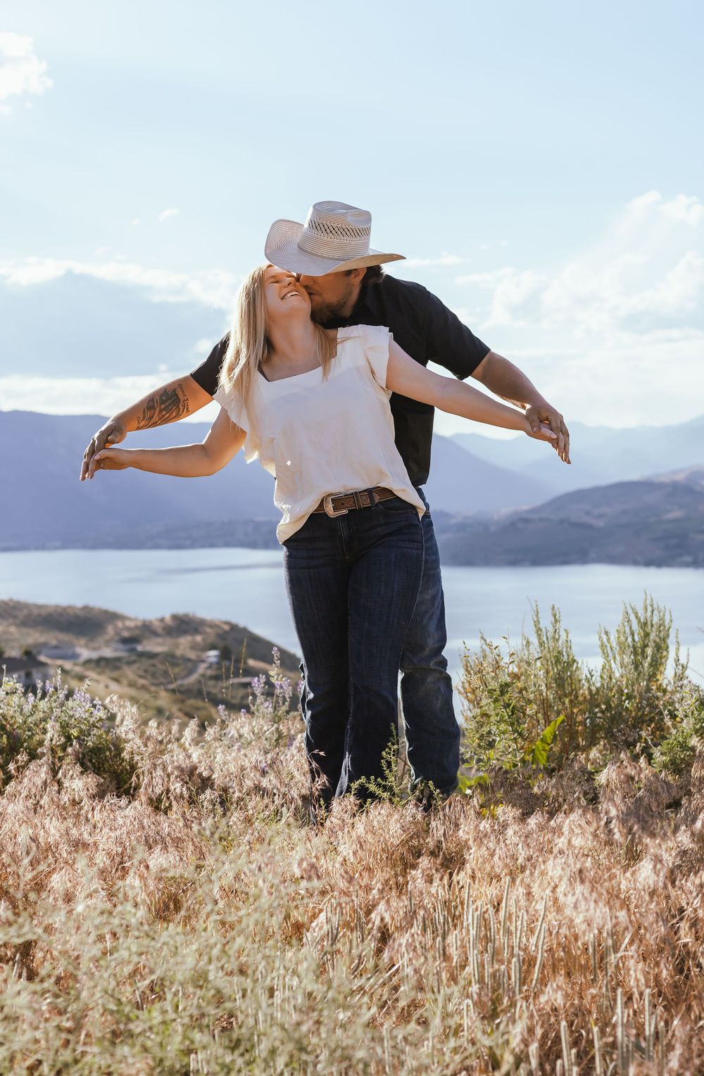 A couple shares a romantic moment on a hilltop overlooking a lake, surrounded by golden grass and mountain views.