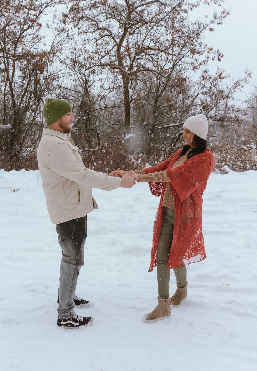 Two people hold hands while standing in a snowy winter landscape with bare trees in the background.