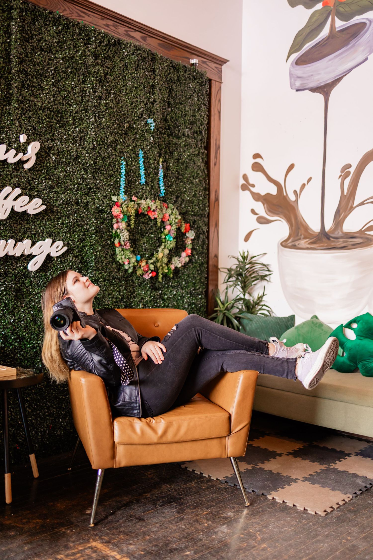 Woman lounges in leather chair against green wall with decorative hanging wreath and coffee signage.