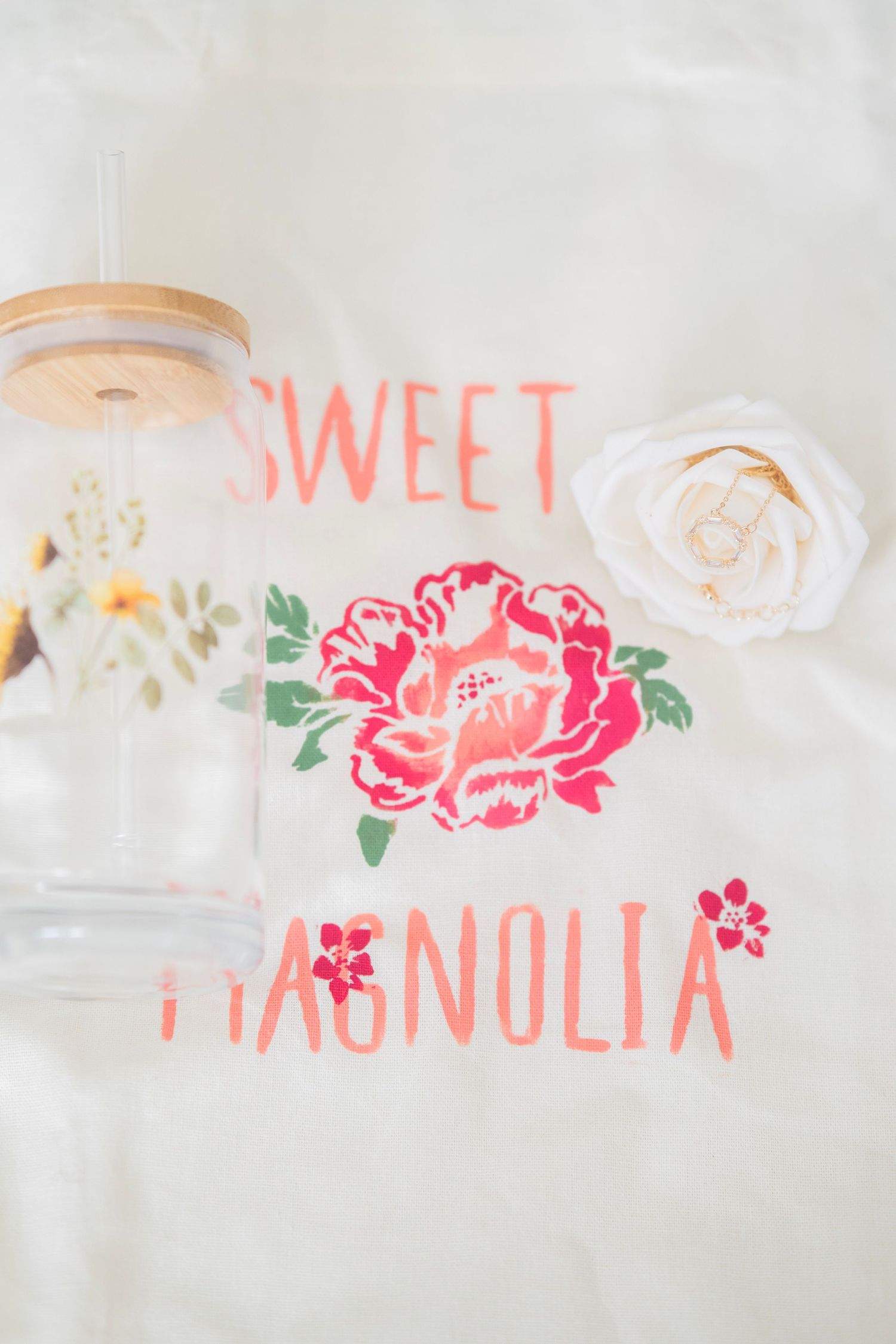 Sweet Magnolia branded tote bag with floral design displayed with a glass tumbler and white rose.