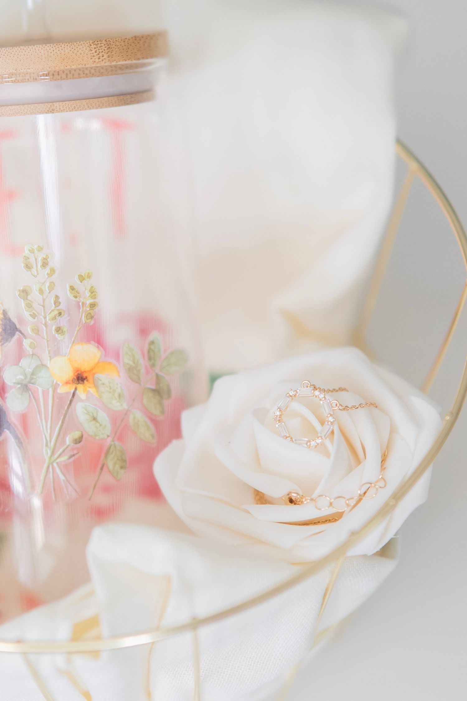 Close-up of floral glass tumbler with gold necklace draped over white rose on decorative tray.