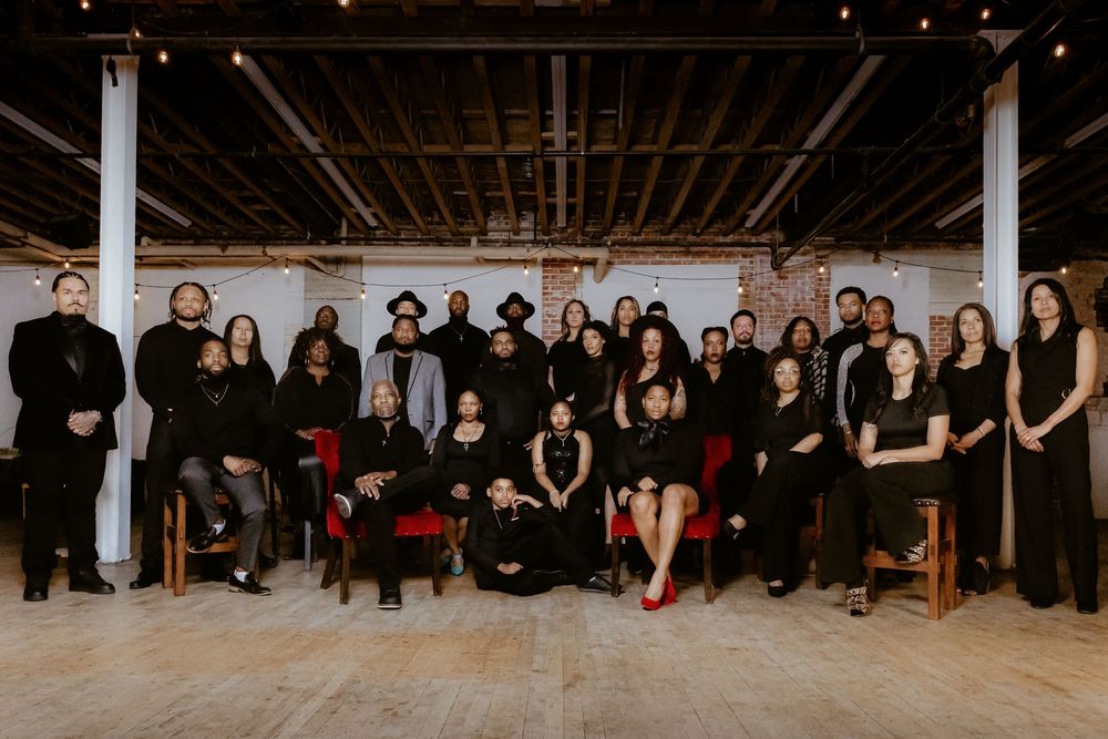 Wide panoramic shot of performers in black attire seated along studio floor.