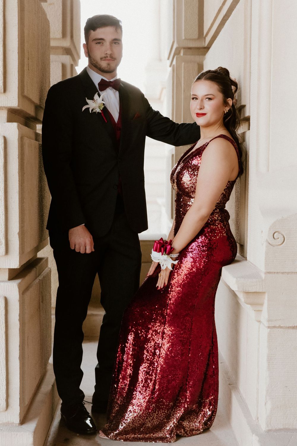 Couple in formal wear poses on elegant white stairs, with woman in sparkling red gown.