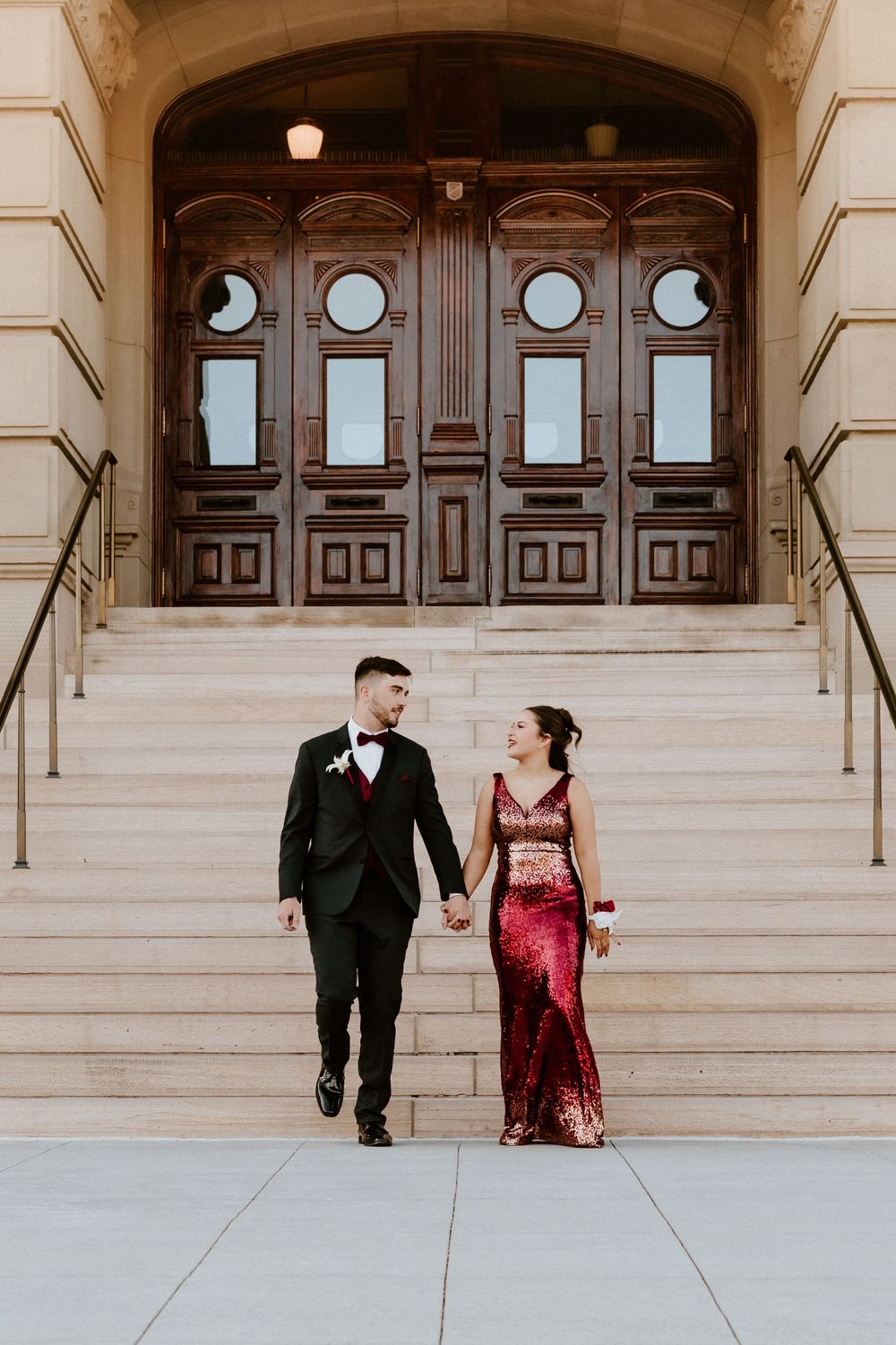 Elegant couple walks down stone steps in front of grand wooden doors at historic building.