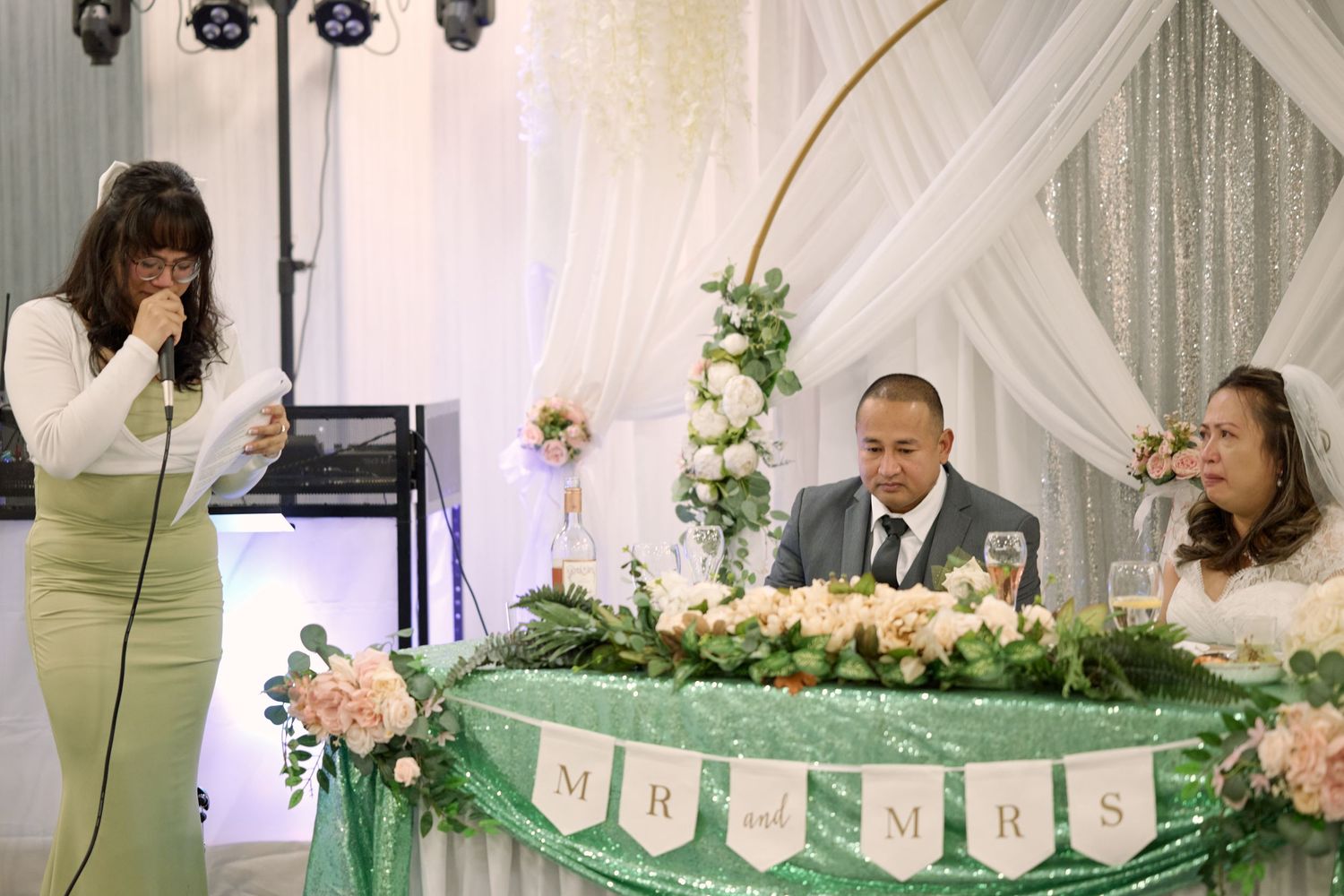 Wedding reception showing event planner speaking at microphone near decorated head table with floral arrangements.
