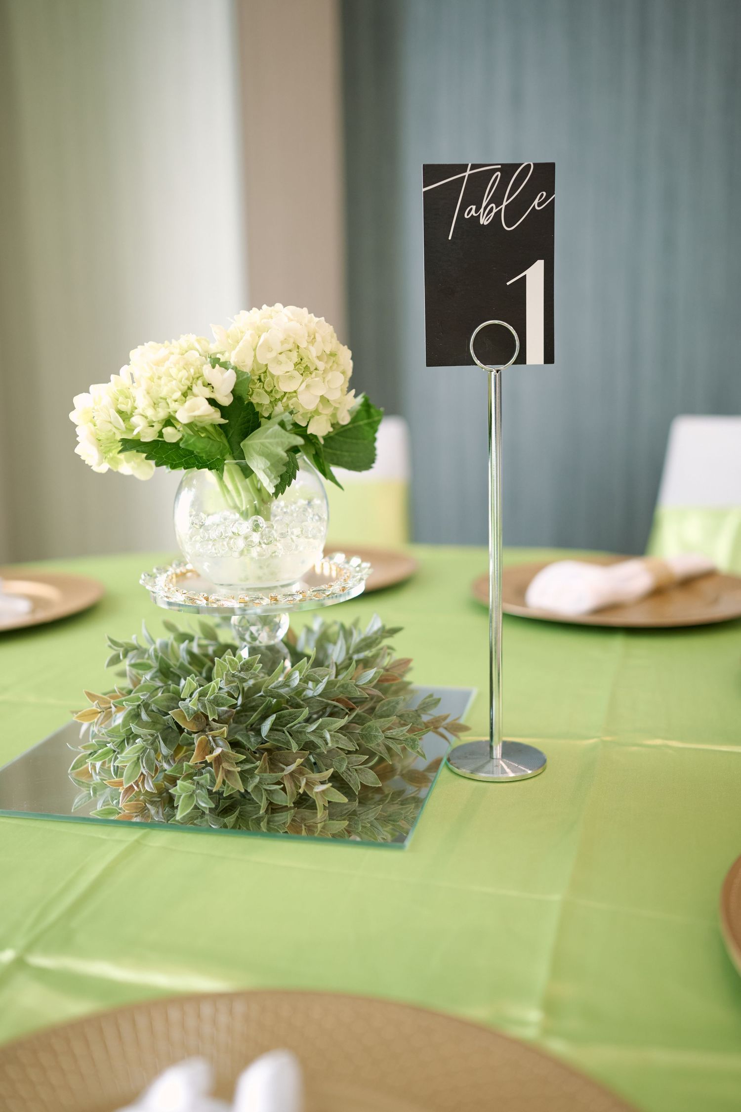Elegant wedding table setting with green tablecloth, white hydrangea centerpiece and black table number one sign.