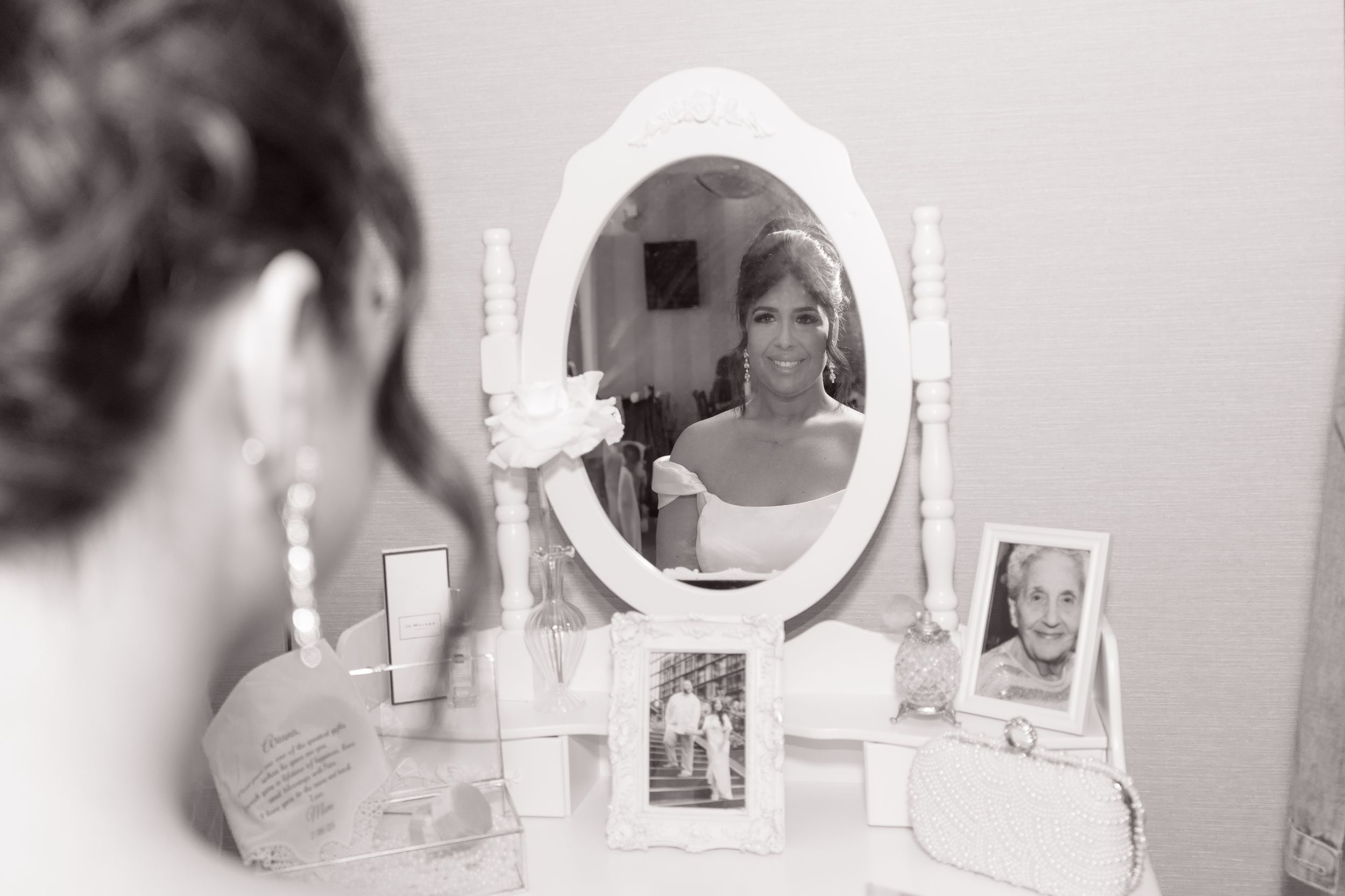 Black and white image showing a reflection in a white vanity mirror with framed photos and beauty products on the surface.