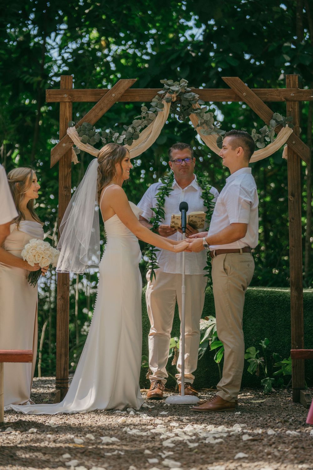 Rustic outdoor wedding ceremony under wooden arch decorated with greenery and flowers in a wooded setting.