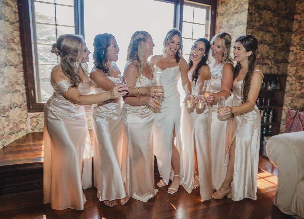 Elegant bridal party in matching champagne silk dresses gather by window in rustic stone-walled venue.