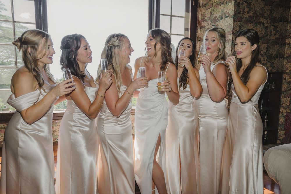 A group of bridesmaids in matching silk dresses share laughter and champagne during a wedding celebration.