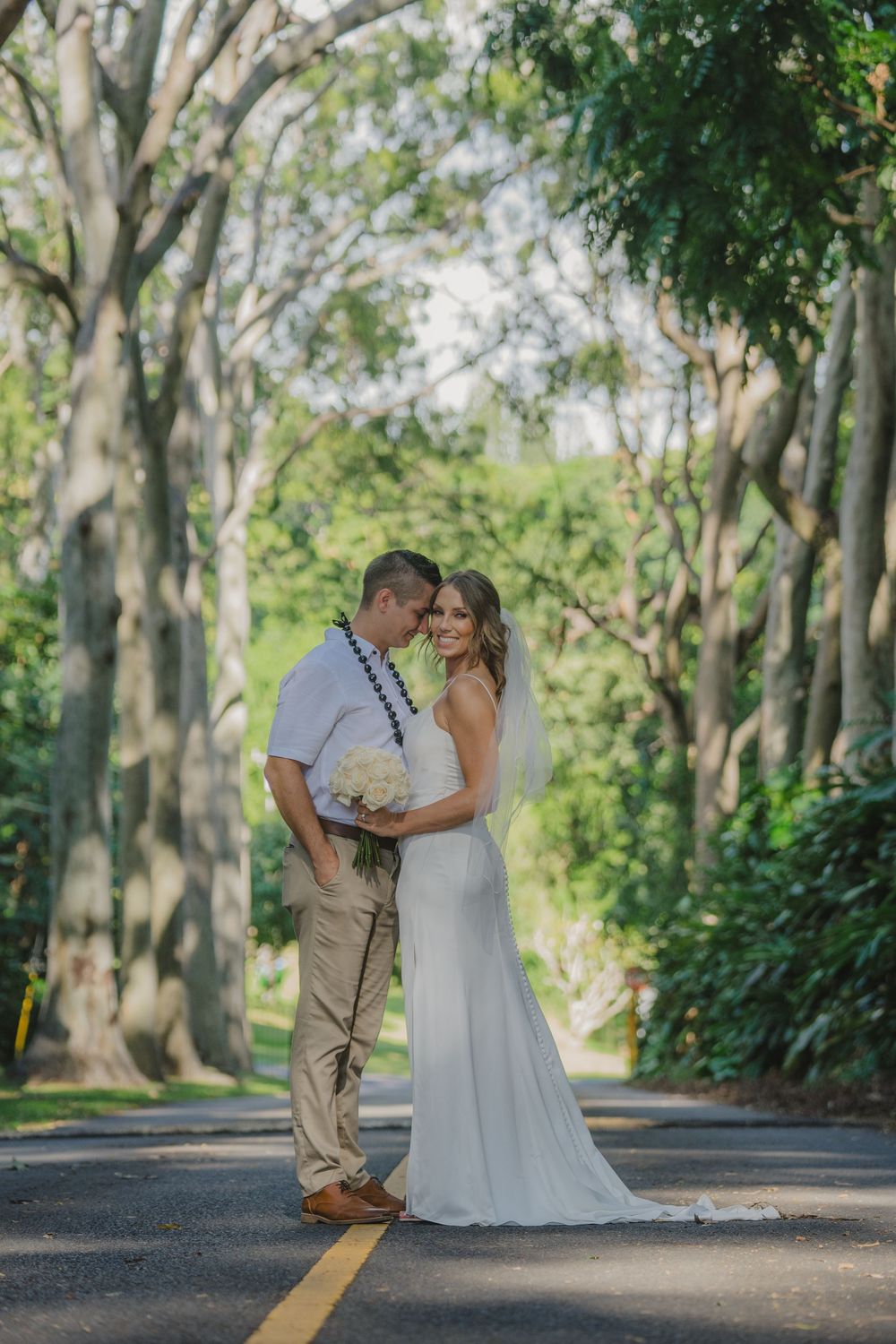 A romantic wedding couple stands embracing on a tree-lined road on their special day.