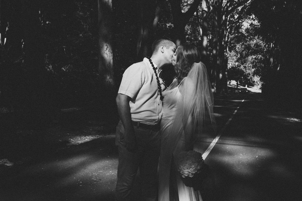 A romantic black and white photo of a couple sharing a kiss on a sunlit path beneath tall trees.