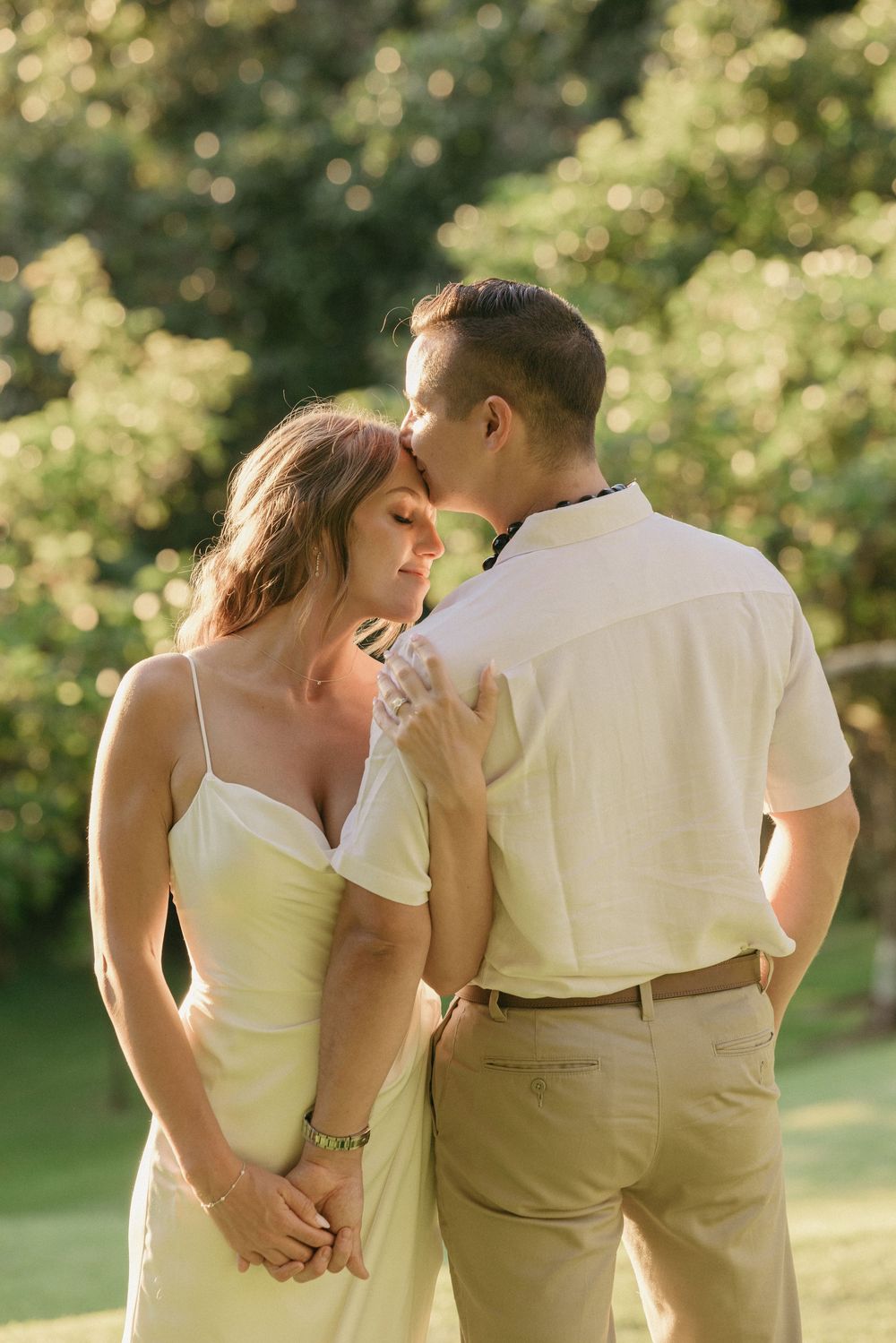 A romantic couple shares an intimate moment outdoors in soft golden light with the woman in white and man in beige attire.