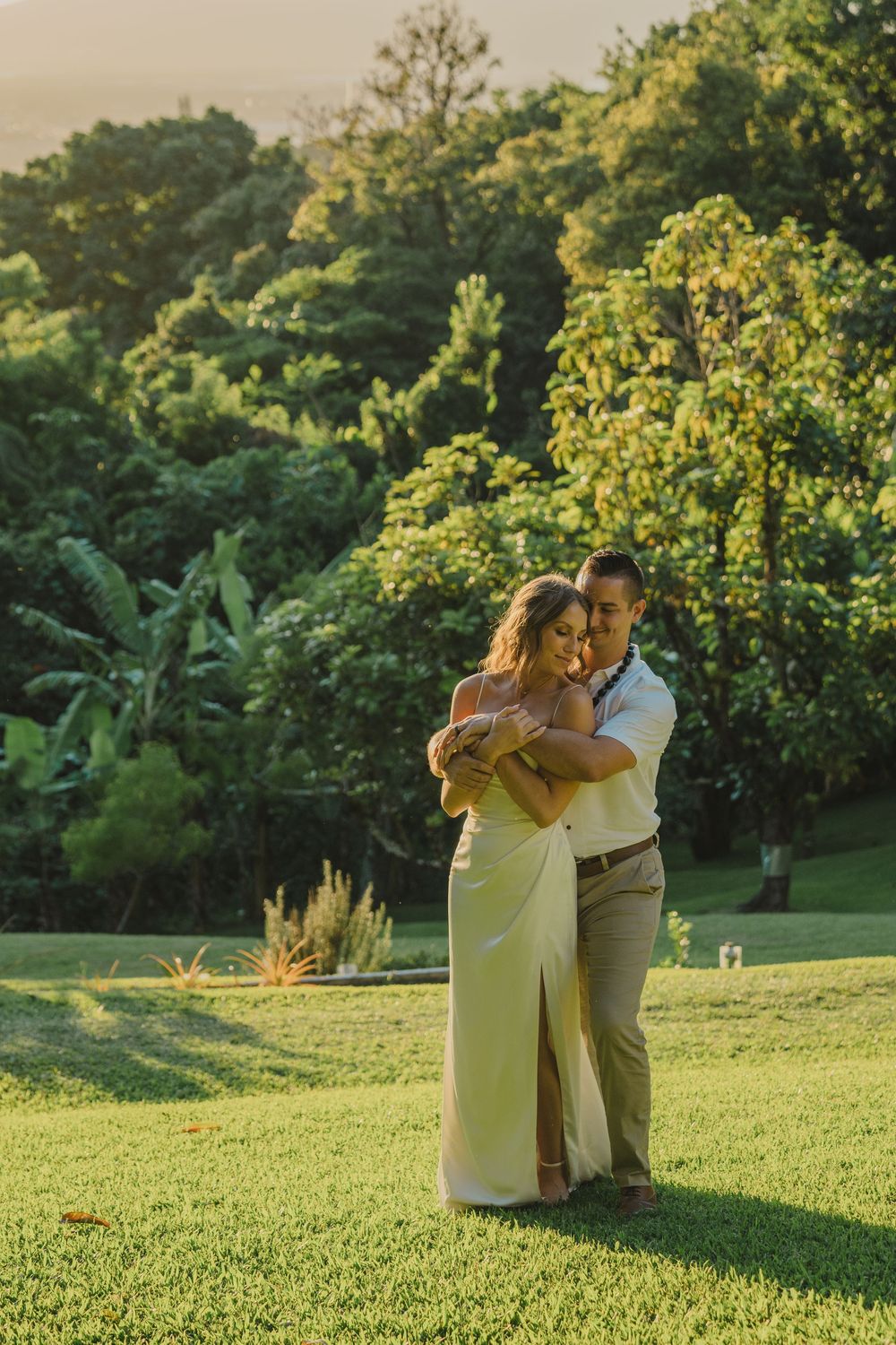 A couple embraces on a lush green lawn at golden hour with tropical foliage and trees in the background.