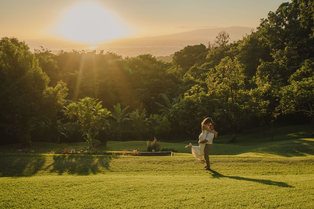 A couple embraces in a scenic grassy field during golden hour sunset with lush green mountains in the background.