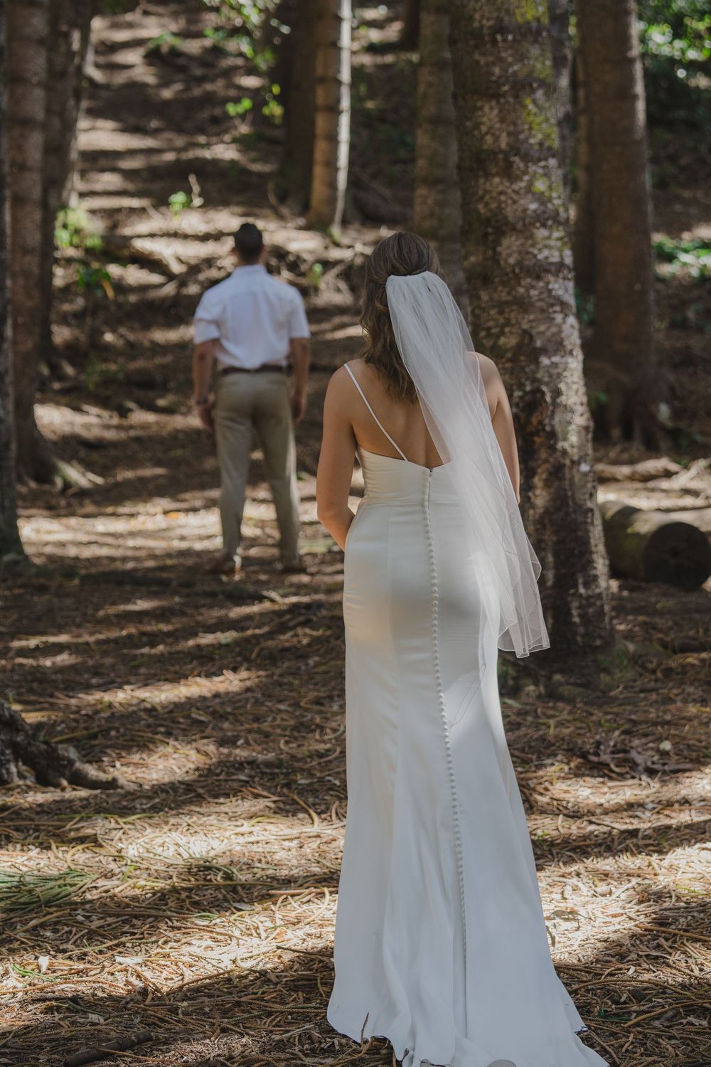 Wedding couple shares a private moment in a sunlit forest setting, the bride wearing an elegant white form-fitting gown and veil.