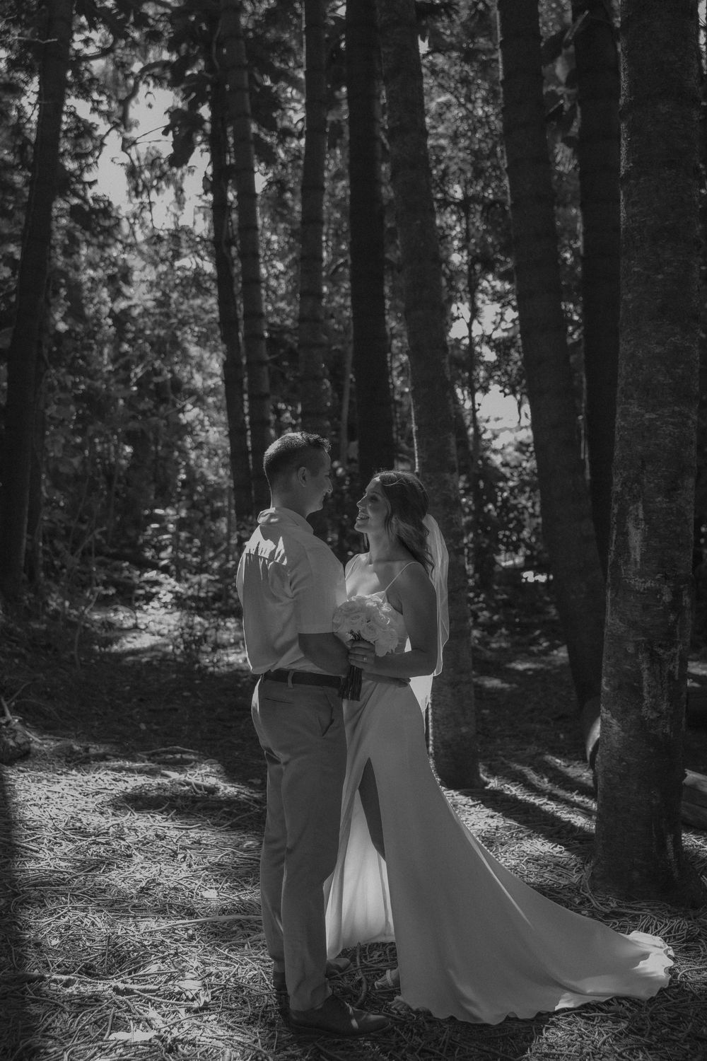 A romantic couple shares an intimate moment in a sunlit forest clearing wearing elegant wedding attire.