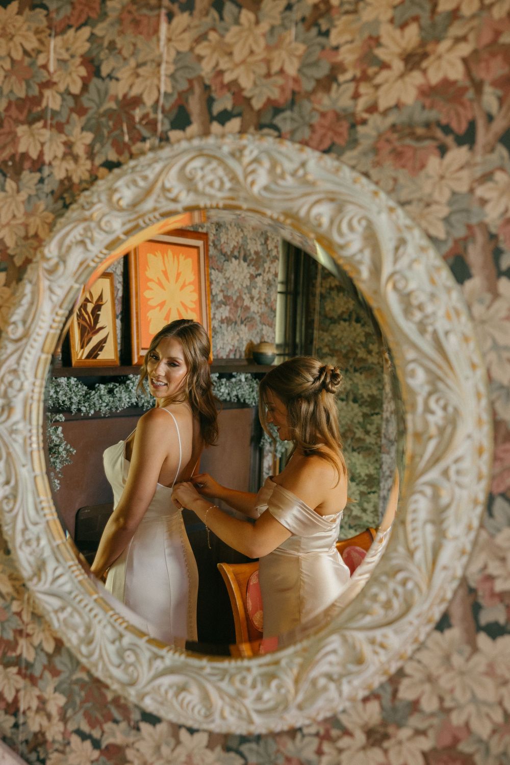 A reflection in an ornate white oval mirror shows wedding preparations in a floral wallpapered room.