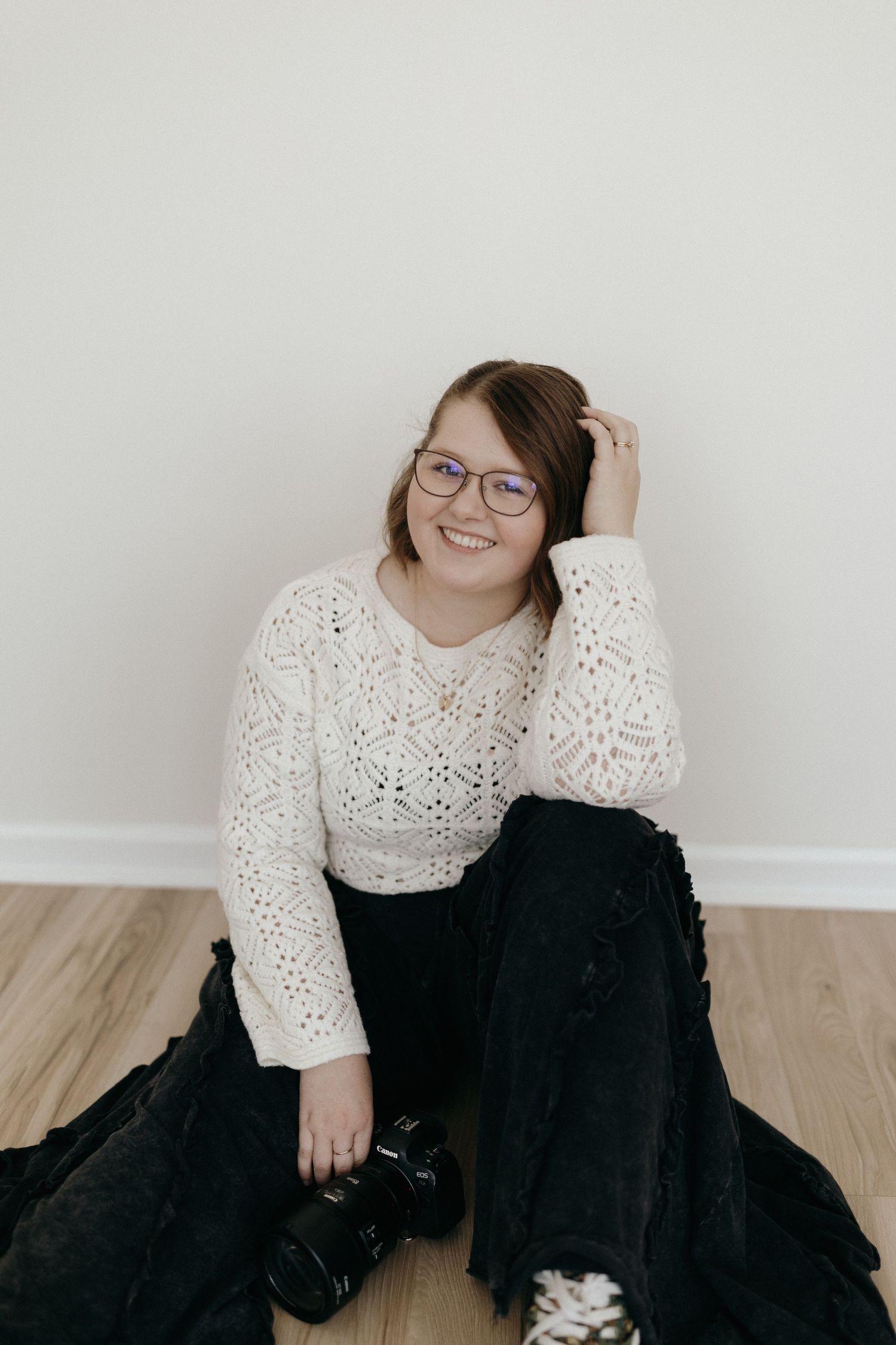 Photographer sitting on hardwood floor wearing white crochet sweater and black pants with camera nearby.