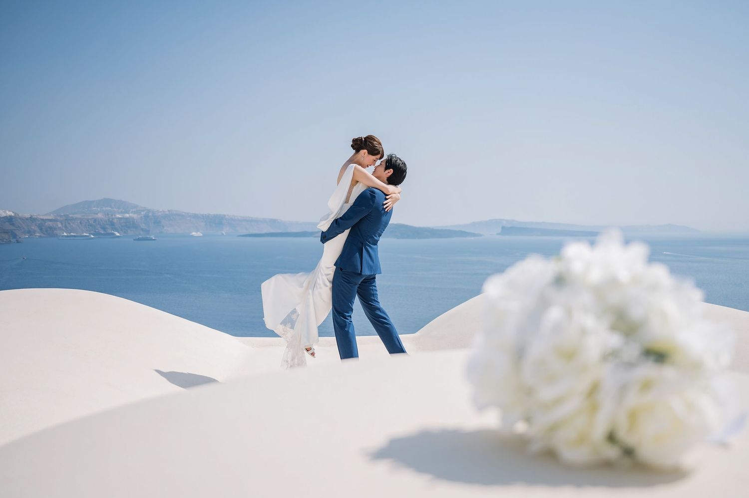 Newlyweds embrace on white rooftop overlooking blue Mediterranean waters with white floral decor.