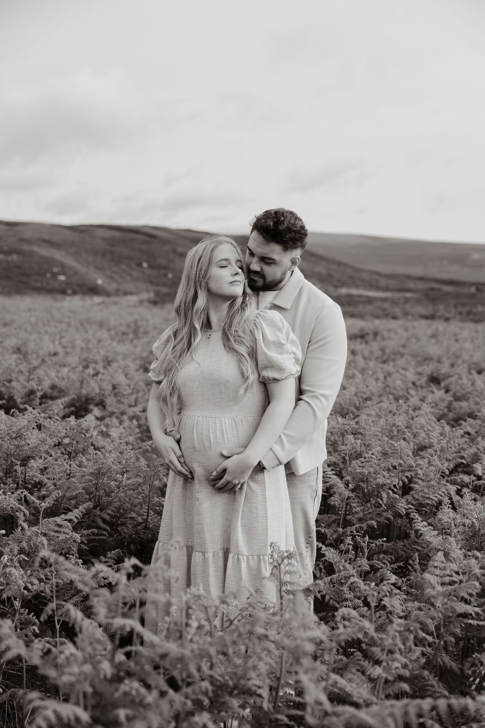 Black and white photo of a couple embracing in a field, woman in floral dress, man holding her from behind.