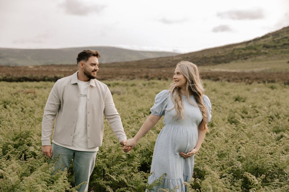 Couple holding hands in a lush green field, woman in blue maternity dress, mountains in background.