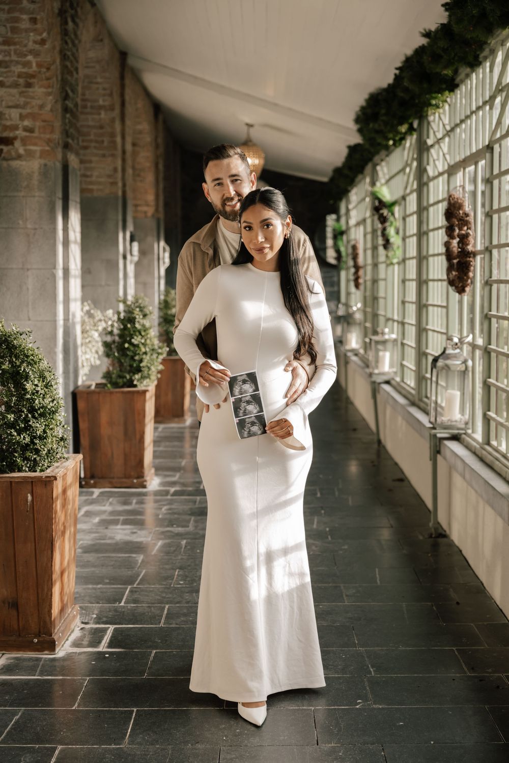 Expecting couple poses in elegant covered walkway with greenery as woman in white dress shows ultrasound photos.