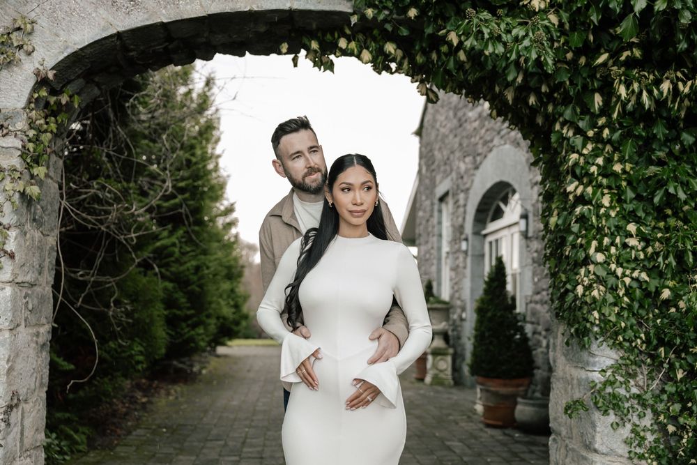 A couple poses under an ivy-covered stone archway with elegant landscaping in a romantic garden setting.
