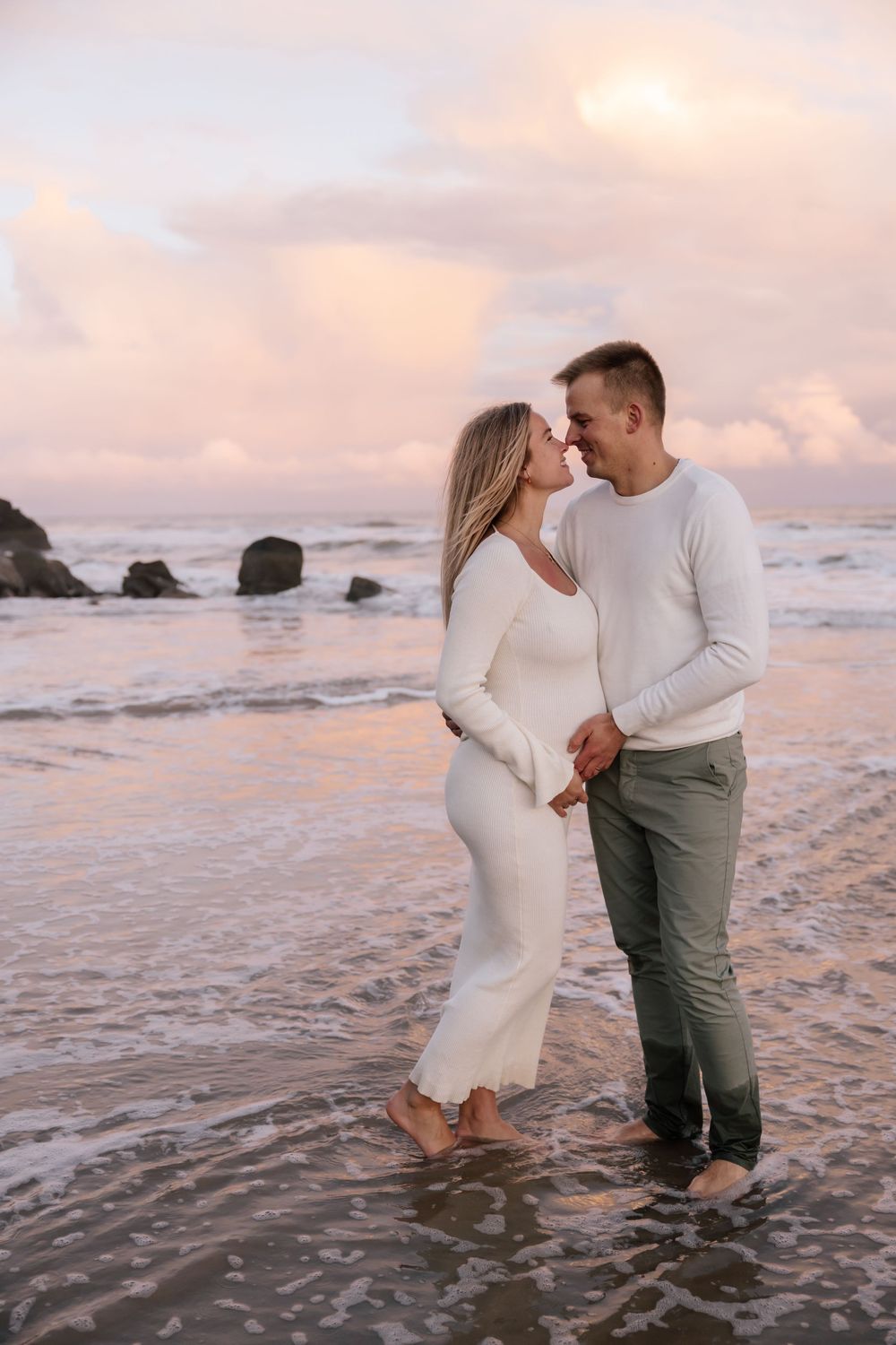 A couple share an intimate moment at sunset on a beach with gentle waves and rocks in the background.