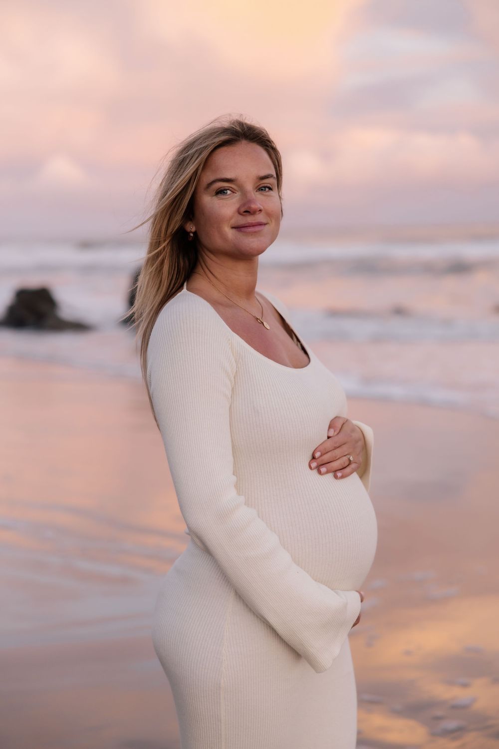 Pregnant person in white dress stands on beach at sunset with warm glowing sky and ocean in background.