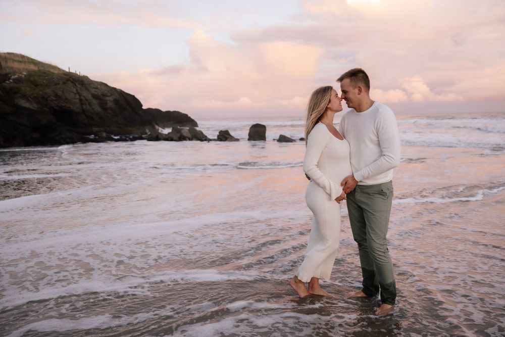 A couple shares a romantic moment on a rocky beach at sunset with waves crashing behind them.