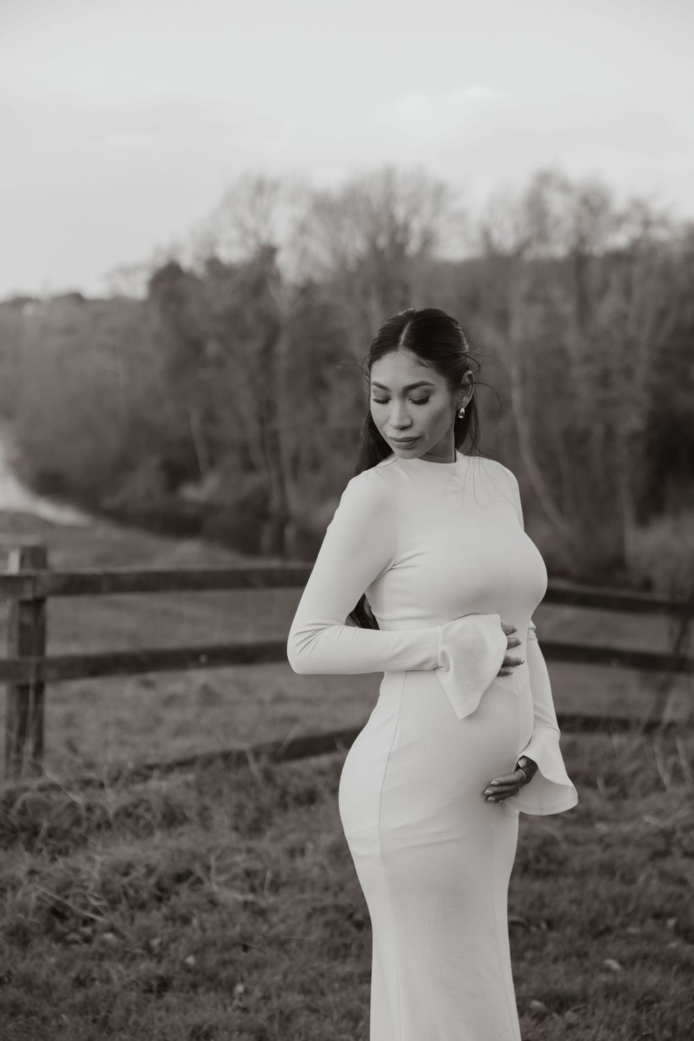 A person in an elegant white long-sleeve gown poses outdoors in a black and white maternity photoshoot.