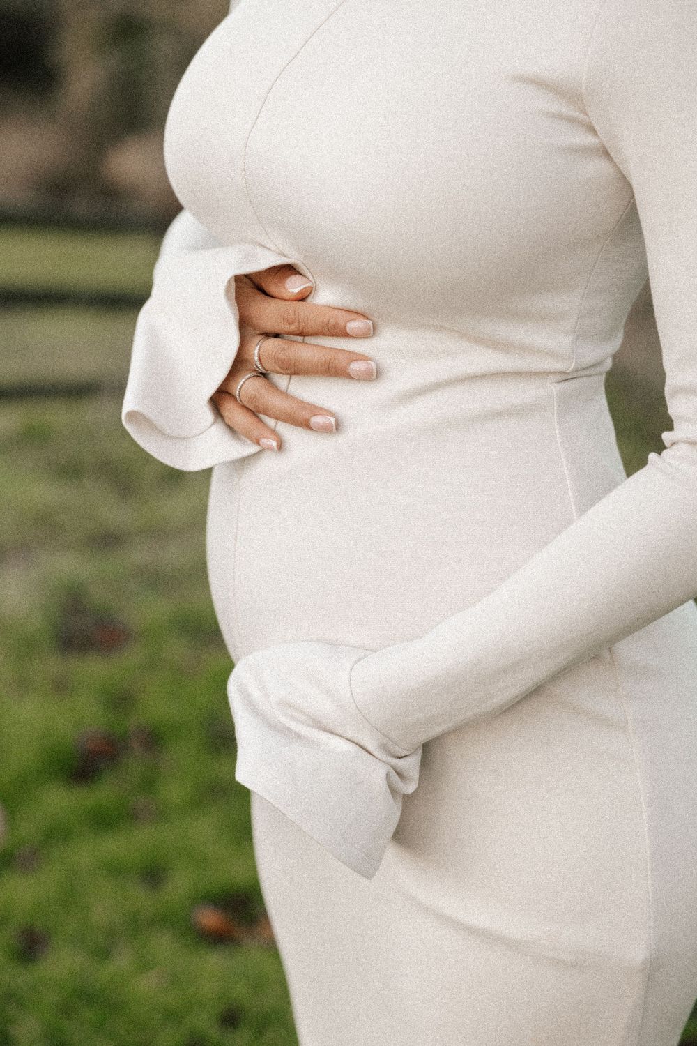 Close-up of pregnant woman in white dress cradling her belly while standing in a garden setting.