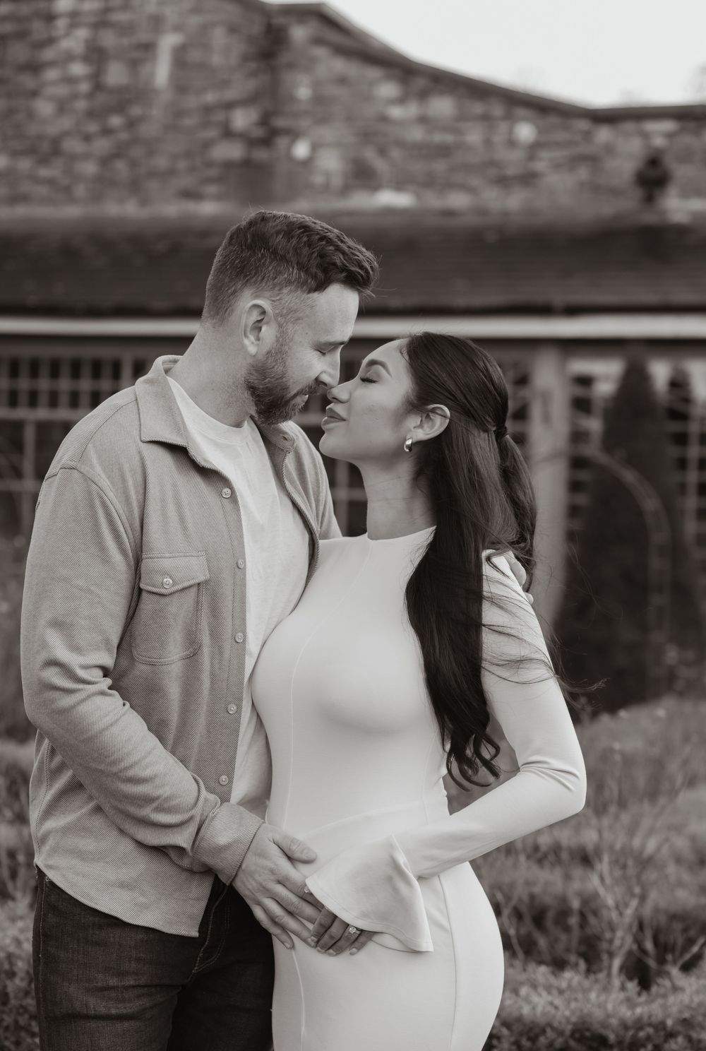 Black and white photo of a romantic couple embracing outdoors in casual attire against a stone wall background.