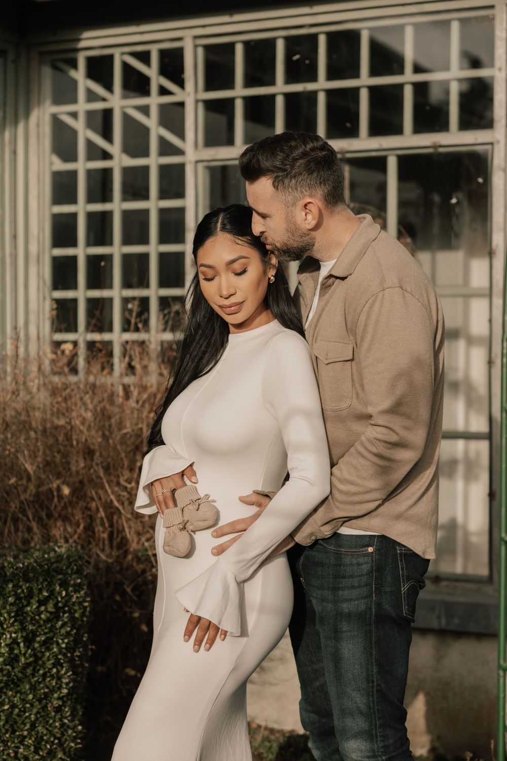 Expecting couple in white dress and tan jacket embrace near window during maternity photo session at sunset.