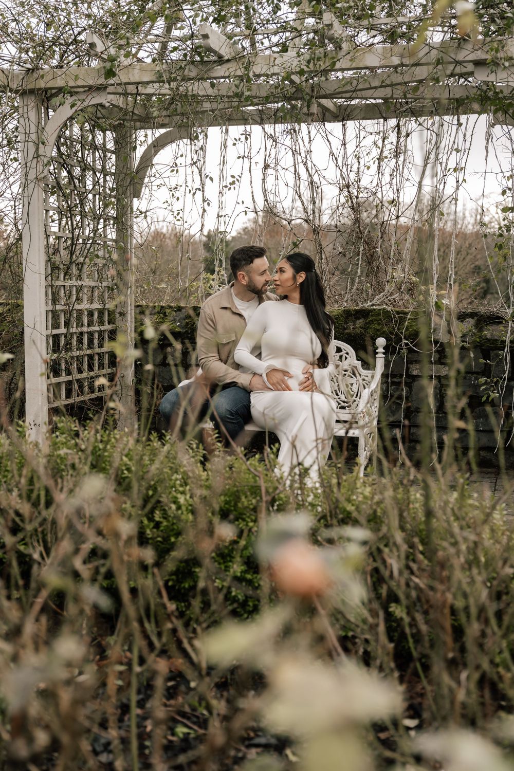 Romantic couple sharing an intimate moment on white bench under wooden garden pergola surrounded by wild foliage.