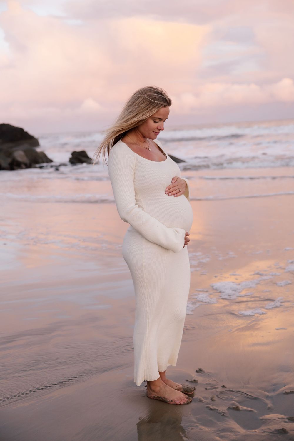 Expectant mother in a flowing white dress stands peacefully on a beach at sunset with waves rolling in the background.