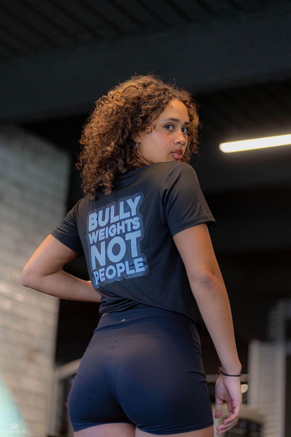Athletic person with curly hair wearing a black t-shirt with 'Bully weights not people' text and dark workout shorts.