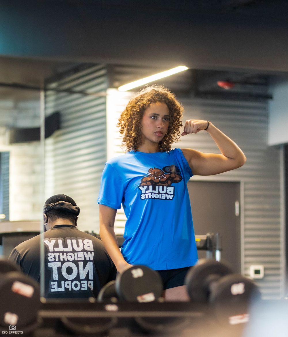 Athletic person in blue t-shirt flexing bicep in front of mirror at fitness facility.