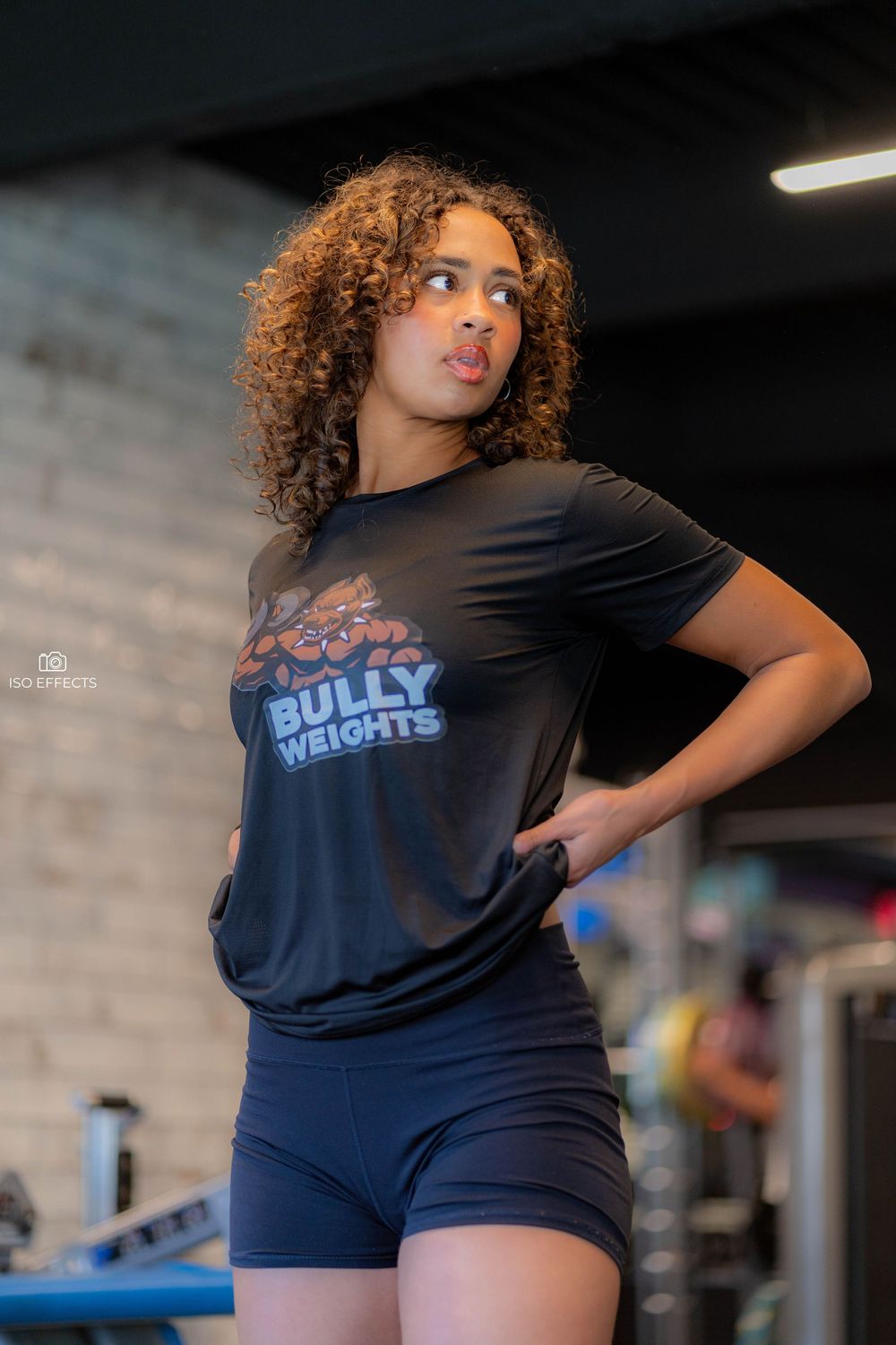 Athletic model wearing Bully Weights branded black t-shirt and navy shorts in gym setting.