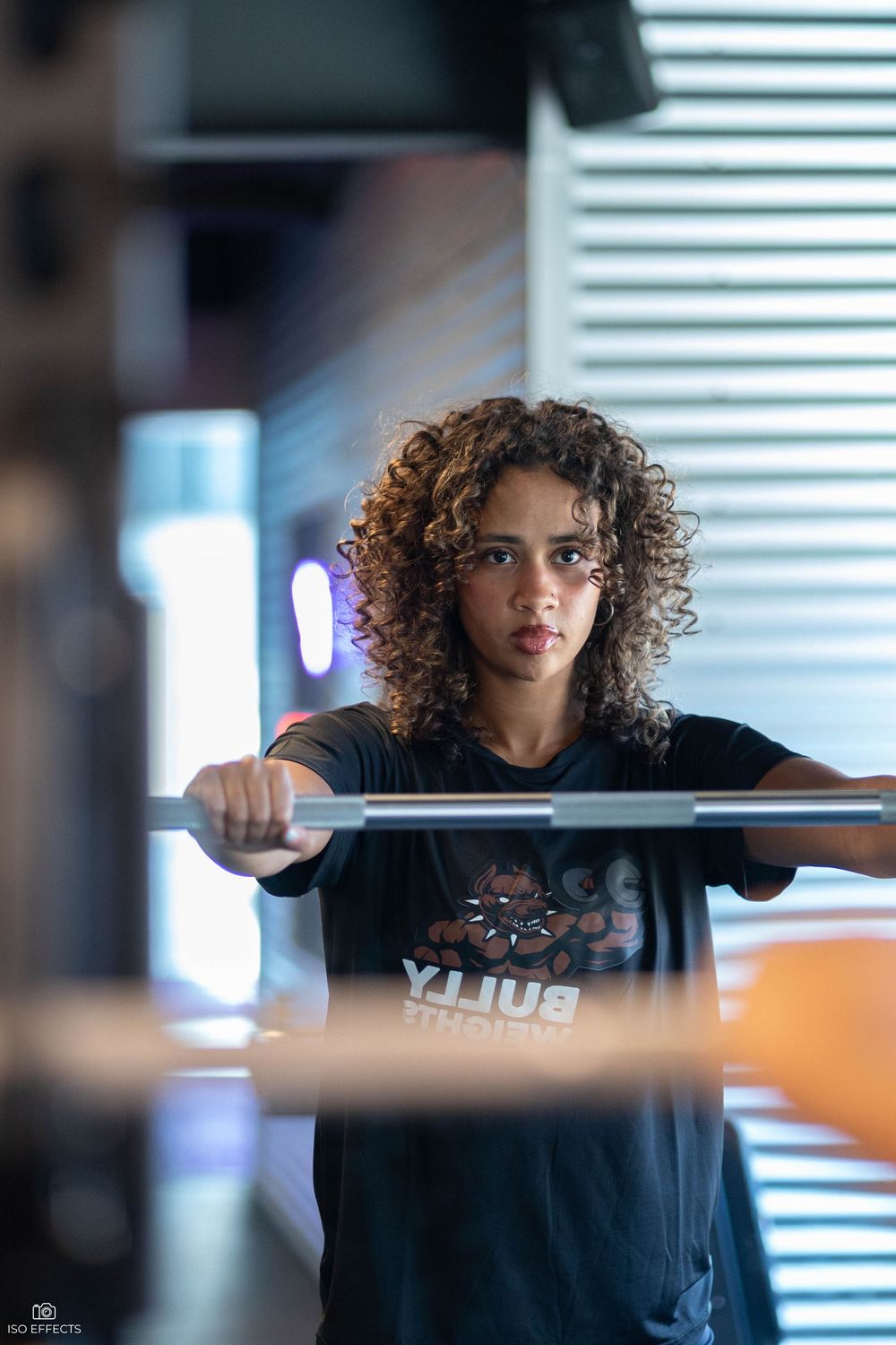 Athletic person wearing a black t-shirt with curly hair performs a weightlifting exercise in a gym with natural lighting.