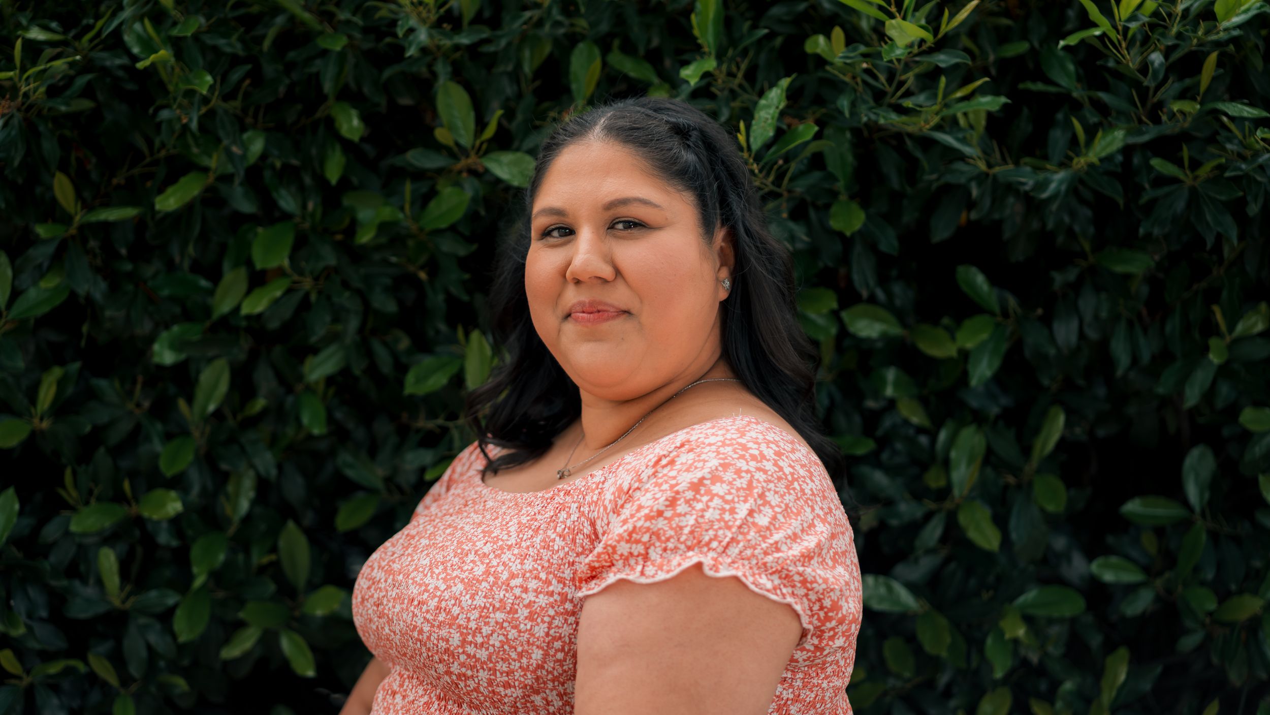 Person wearing peach-colored lace top smiles while standing in front of green leafy background.