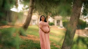 Expecting mother in a flowing pink dress stands in a dreamy outdoor setting with trees and sunlight.