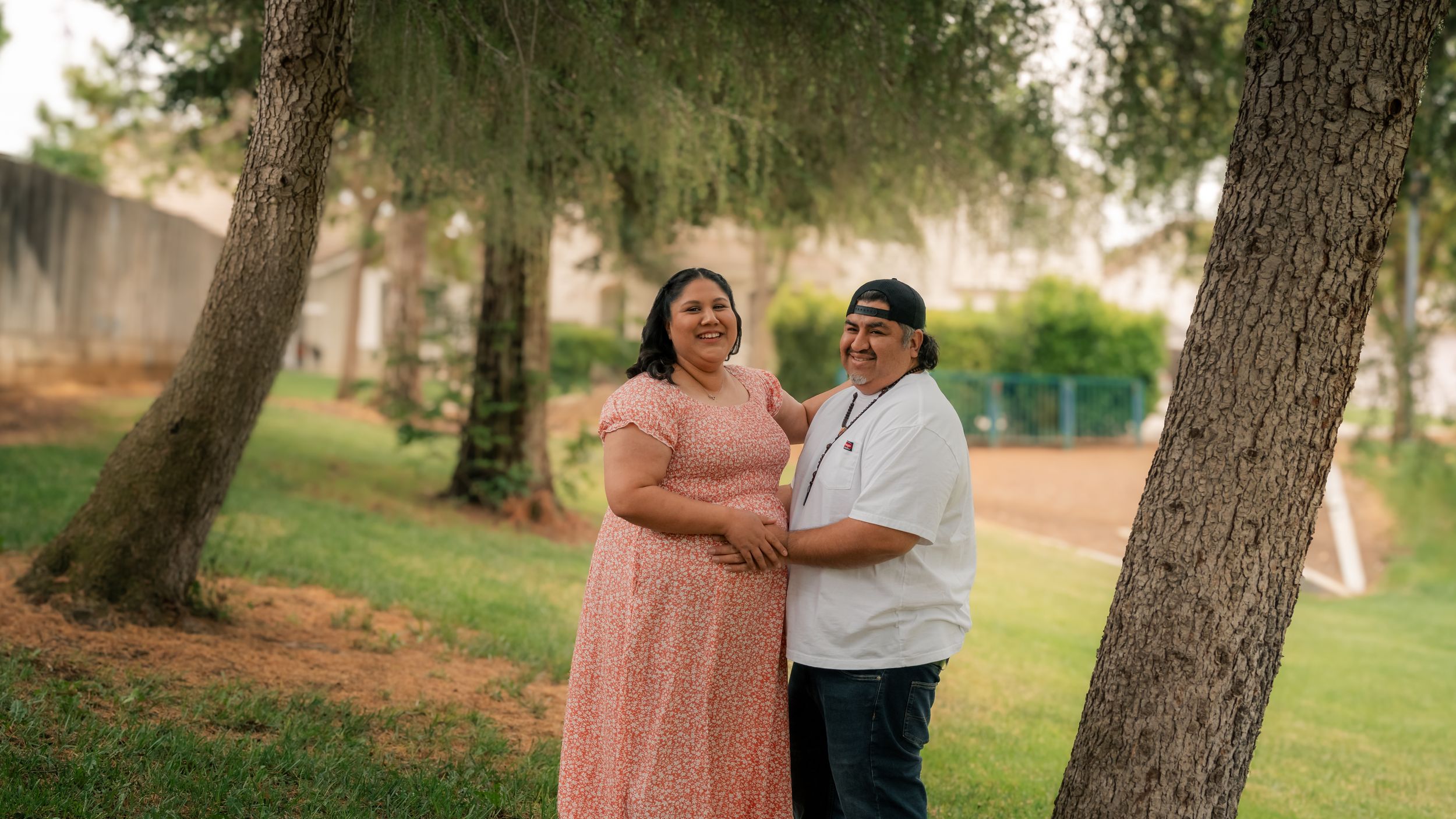 A couple embraces outdoors in a grassy park area with pine trees creating a warm, intimate moment together.