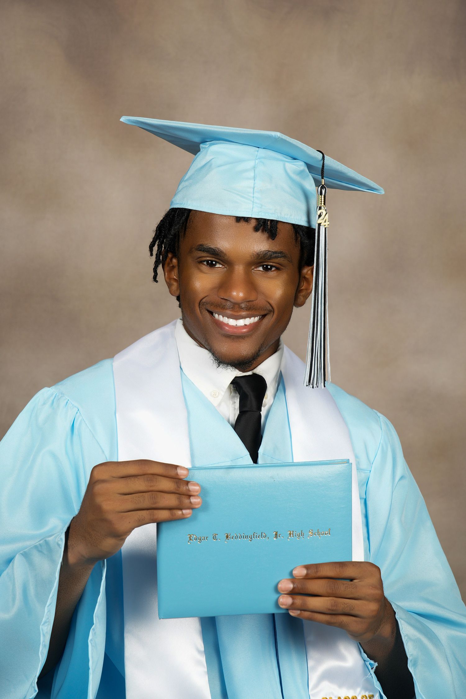 A graduate wearing light blue cap and gown with white sash holds a diploma during portrait session.