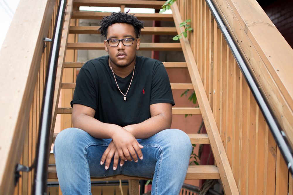 A person wearing glasses and a black t-shirt sits on wooden stairs in a casual outdoor portrait.